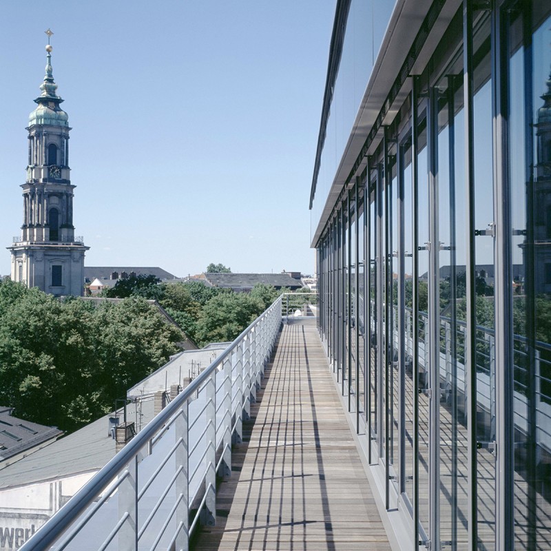 Terrasse eines Gebäudes mit vielen Fenstern mit Blick auf Bäume und einen Turm